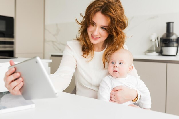 Portrait of smiling woman sitting and holding her cute little baby in hand while happily using her tablet