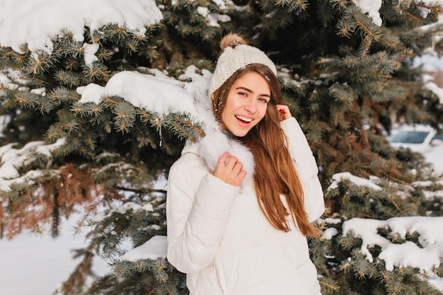 Free Photo portrait of smiling woman in warm white coat posing beside tree in frosty day. outdoor photo of romantic lady with long hair standing in front of snowy spruce during winter photoshoot.