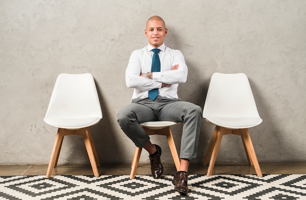 Free photo portrait of a smiling young businessman sitting on chair with his arm crossed