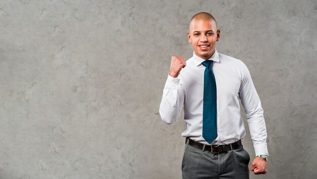 Portrait of a smiling young businessman standing against grey wall clenching his fist