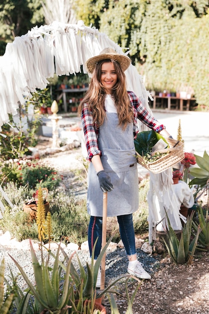 Free Photo portrait of a smiling young female gardener holding hoe and basket in the garden