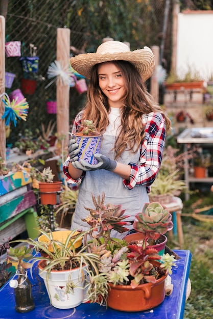 Free photo portrait of a smiling young female gardener wearing hat holding painted pot plant in greenhouse