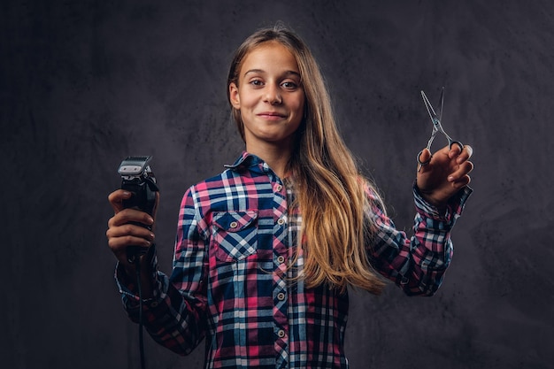 Portrait of a smiling young girl dressed in shirt holds trimmer and scissors. Isolated on a dark textured background.