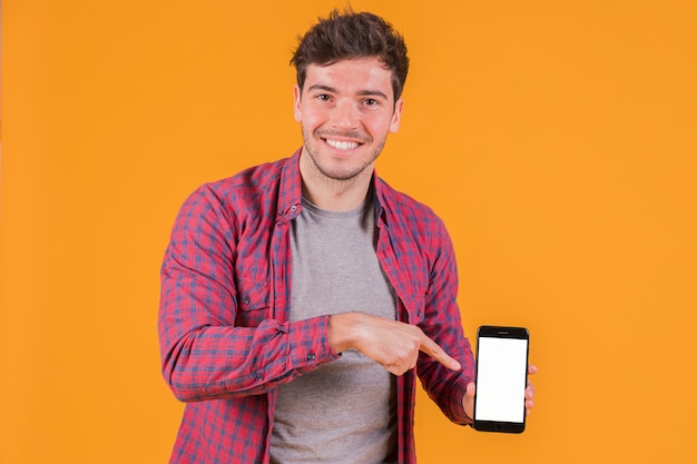 Free photo portrait of a smiling young man pointing his finger on mobile phone against an orange background