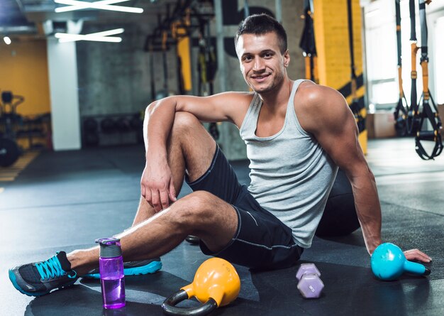 Portrait of a smiling young man sitting on floor near exercise equipments in gym