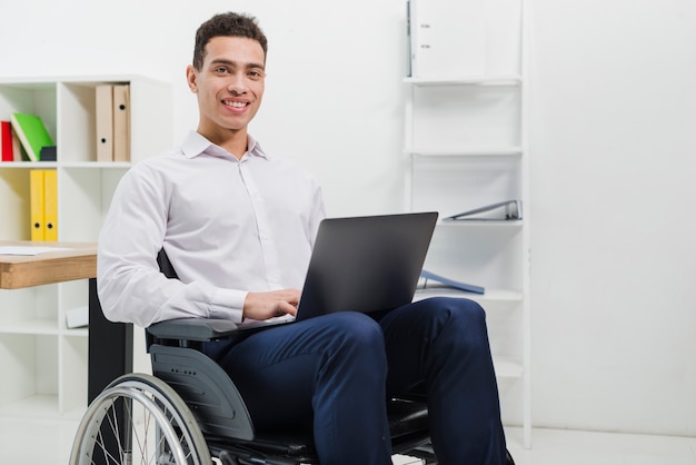 Portrait of a smiling young man sitting on wheelchair with laptop looking at camera