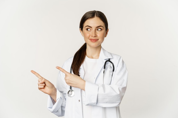 Portrait of smiling young woman doctor healthcare medical worker pointing fingers left showing clini...