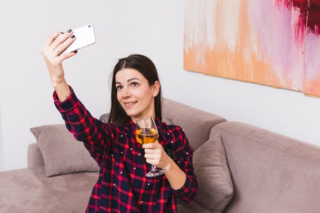 Portrait of a smiling young woman holding wineglass taking selfie on mobile phone at home