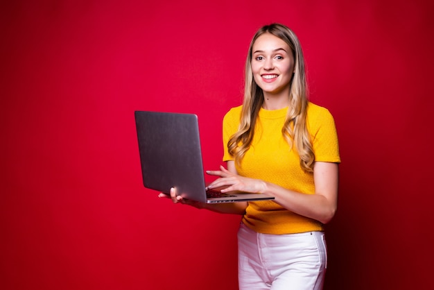 Portrait of smiling young woman holding, working on laptop pc computer isolated on red wall.