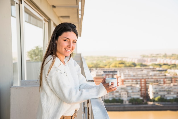 Free photo portrait of a smiling young woman standing in balcony holding white coffee cup