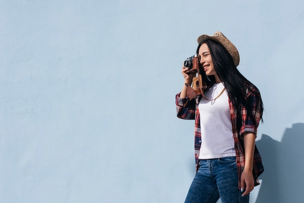 Free photo portrait of smiling young woman taking picture with camera standing near blue wall