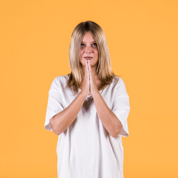 Portrait of smiling young woman with praying gesture standing against yellow wall