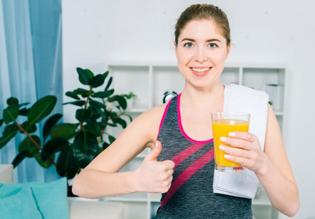 Portrait of a smiling young woman with white towel over her shoulder holding glass juice showing thumb up sign