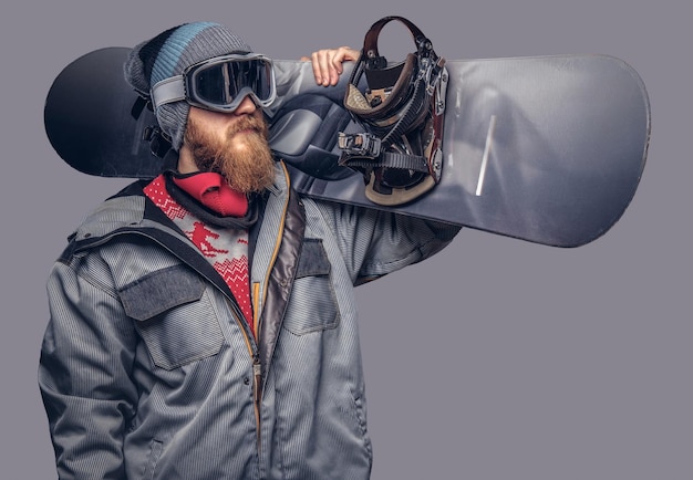 Portrait of a snowboarder dressed in a full protective gear for extream snowboarding posing with a snowboard on his shoulder at a studio. Isolated on a gray background.