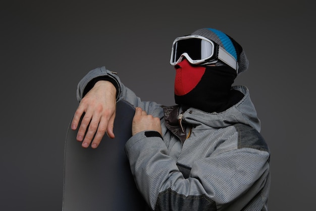 Free photo portrait of a snowboarder wearing full protective equipment for extreme snowboarding posing with snowboard at a studio, isolated on a dark background.