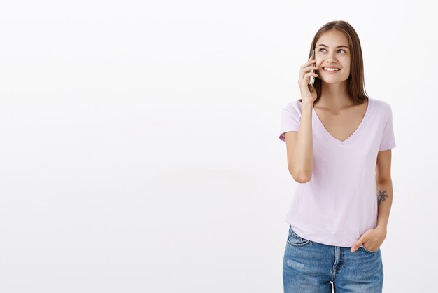 Portrait of socisble happy and carefree confident young woman with tattoo in white t-shirt smiling delighted gazing up while holding smartphone near ear talking via cellphone with friend