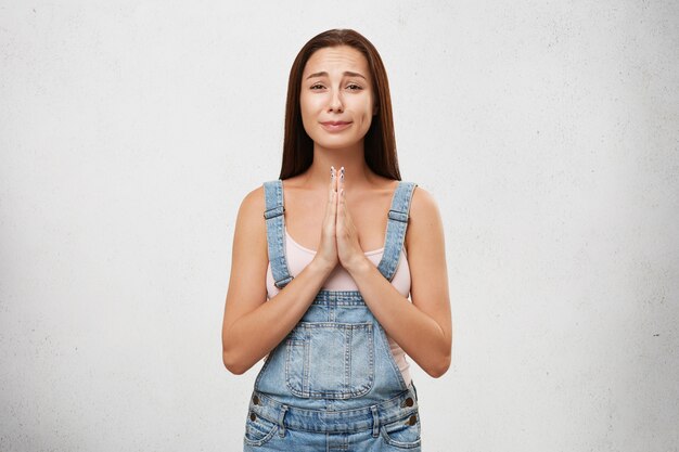 Portrait of sorrorful female wearing white shirt and denim overalls, keeping palms together, going to cry while asking for forgiveness. Young brunette woman with sullen look isolated over white wall