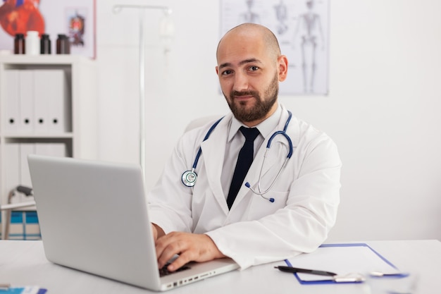 Portrait of specialist physician man looking into camera working in meeting conference room analyzing sickness expertise using laptop computer. Doctor prescribing pills medication healthcare treatment