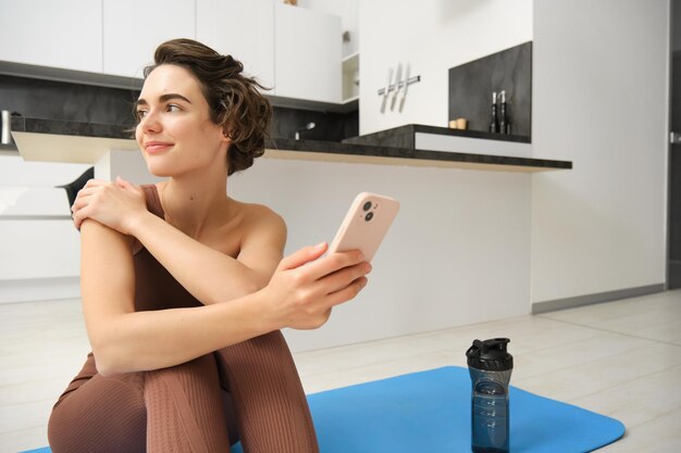 Portrait of sporty girl with mobile phone sitting on yoga mat using smartphone during workout at hom