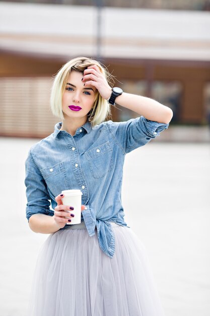 A portrait of a standing dreamy blonde girl with bright pink lips hoding a cup of cofee and holding one hand near her head