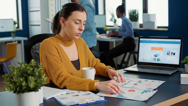 Free photo portrait of startup employee comparing two sheets of paper with charts analyzing financial data while drinking morning coffee. woman working in busy marketing department sitting at desk with laptop.