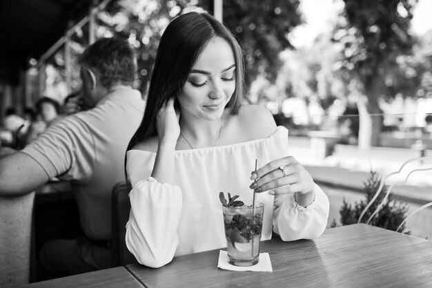 Free photo portrait of a stunning young woman posing with mojito cocktail in cafe next to the park black and white photo