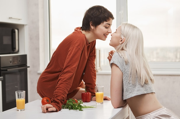 Free Photo portrait of stylish and attractive lesbian in red pullover sitting at kitchen table while kissing girlfriend and smiling happily during breakfast. girl finally found her soulmate