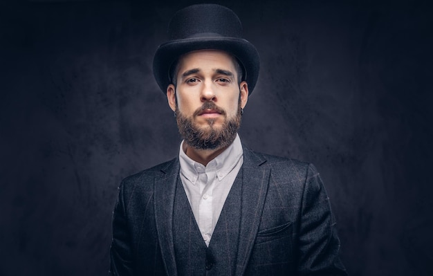 Portrait of a stylish bearded male in an elegant suit and cylinder hat, looking at a camera over dark background.