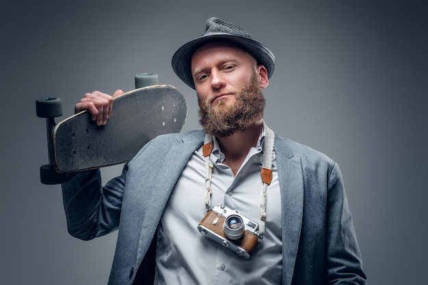 Free photo portrait of a stylish bearded male in a suit and felt hat holds skateboard and vintage slr photo camera in the grey vignette background.