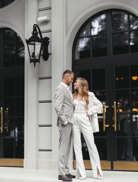 Portrait of stylish brides couple man and woman in white wedding suit looking at each other Posing near modern building restaurant Modern wedding Happy married couple Marriage