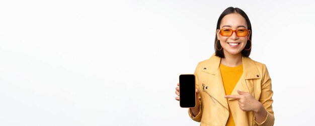 Portrait of stylish korean girl in sunglasses smiling pointing finger at smartphone screen showing mobile phone application standing over white background