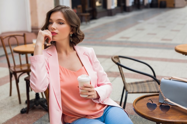 Free photo portrait of stylish romantic woman sitting in cafe drinking coffee, wearing pink jacket and blouse, color trends in apparel, spring summer fashion, accessories sunglasses and bag, thinking, looking