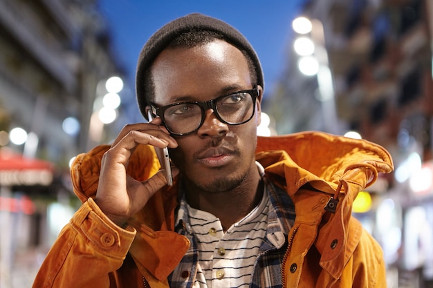 Portrait of stylish young Afro American male having nice conversation on mobile, spending evening outdoors with city lights