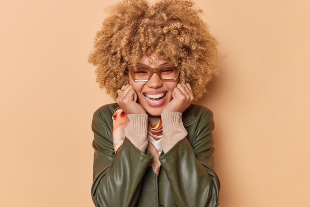 Portrait of stylish young woman keeps hands on face smiles positively looks sincere at camera wears spectacles and leather jacket isolated over brown background hears great news. Happiess concept