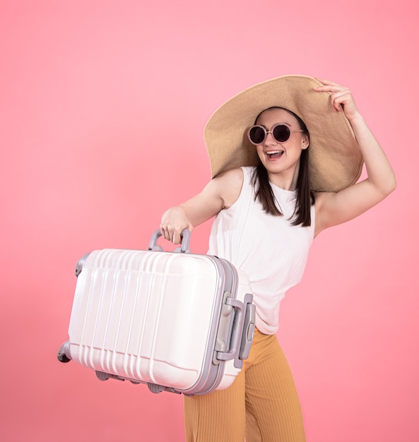 Portrait of a stylish young woman in summer clothes and a wicker hat with a suitcase onn isolated pink