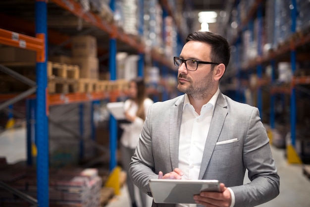 Free photo portrait of successful businessman manager ceo holding tablet and walking through warehouse storage area looking towards shelves