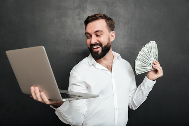 Free photo portrait of successful businessman in white shirt holding fan of money dollar banknotes and silver notebook in both hands over dark gray