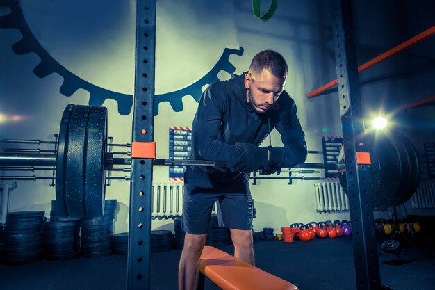 Portrait of super fit muscular young man working out in gym with barbell on blue