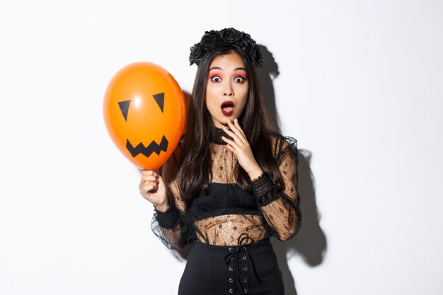 Portrait of surprised asian woman in halloween costume, dressed-up as witch, holding orange balloon with scary face.