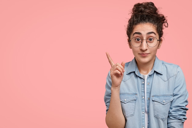 Portrait of surprised brunette Caucasian female looks with puzzled expression, raises fore finger at left upper corner against a pink wall with copy space