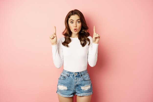 Portrait of surprised caucasian woman, stylish girl pointing fingers up and looking impressed, showing store logo, discount advertisement, pink background