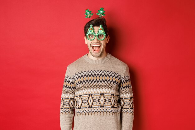 Portrait of surprised and excited handsome guy in party glasses and winter sweater, celebrating christmas and having fun, standing against red background