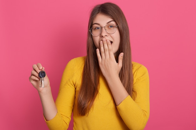 Portrait of surprised female dresses yellow shirt and eyeglasses, holding keys in hands, covering her mouth with hand