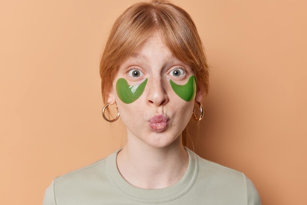 Free Photo portrait of surprised ginger girl keeps lips folded applies green collagen patches to reduce puffiness under eyes wears earrings poses against brown background women skin care and beauty concept