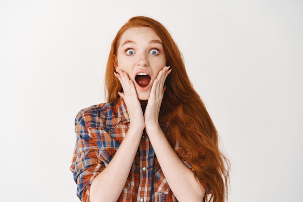 Free photo portrait of surprised girl with long red hair drop jaw of amazement, touching cheeks, standing over white bacground