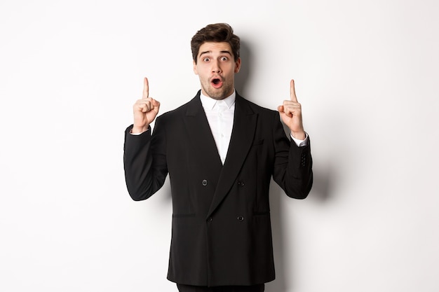 Free photo portrait of surprised handsome businessman in black suit, saying wow and looking amazed, pointing fingers up at copy space, standing over white background