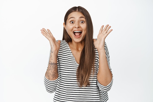 Portrait of surprised and happy woman reacts to big news, shaking hands near face and shouting from joy on white