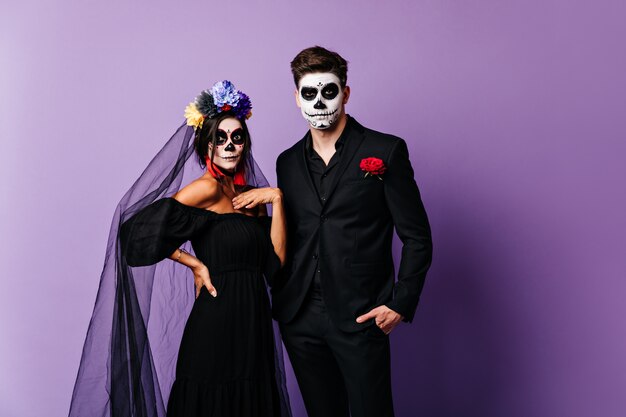 Portrait of surprised lady in image of bride for Halloween and her boyfriend in classic costume with painted face in shape of skull.