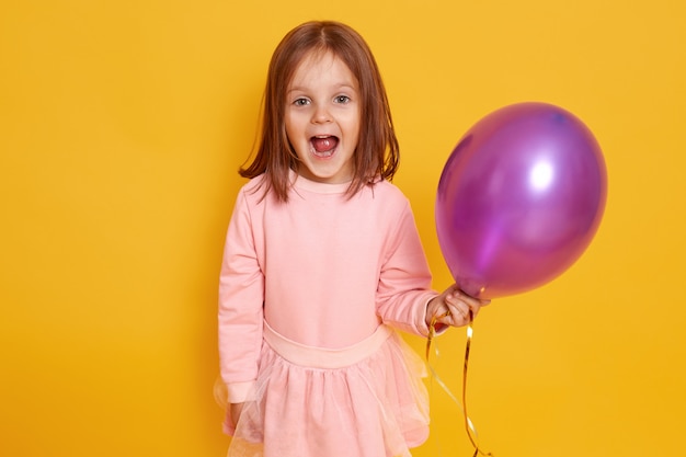 Free photo portrait of surprised little girl with dark straight hair standing over yellow studio beautiful clothes, holding purple ballon in hands