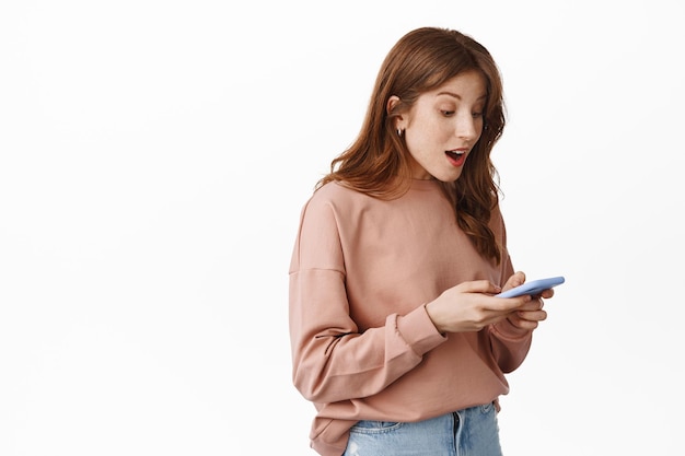 Free Photo portrait of surprised redhead girl looking at phone screen with happy face, reading message notification on mobile, chatting or shopping online, standing over white background